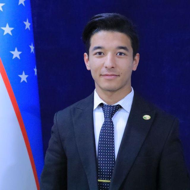 Young Central Asian man with a suit and tie and short hair standing in front of a flag and a blue photo background. 