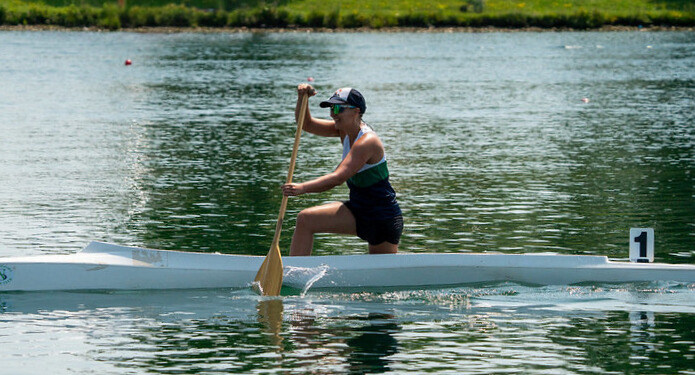 Tarzan paddles in a white, pencil-thin racing canoe with one knee up and one knee down (like a marriage proposal), holding a wooden paddle halfway in the water. There’s a number 1 on the back of her boat. She wears a striped singlet in her team colours, white, green and blue, baseball hat and sunglasses. Her rippling arm muscles are visible. 