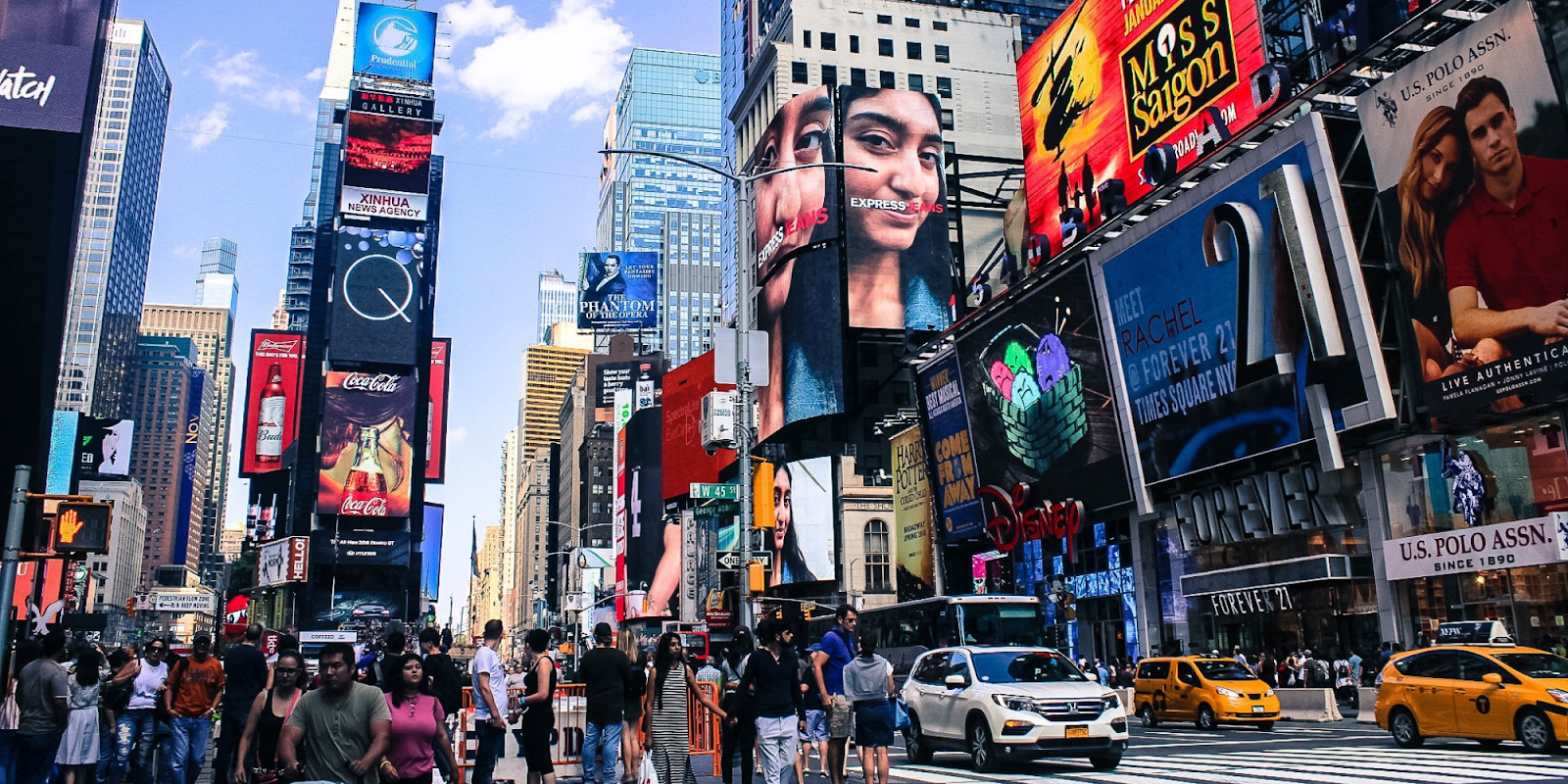 A bustling view of Times Square in New York City, filled with crowds of people and vibrant billboards advertising various brands and shows. Yellow taxis and other vehicles navigate the busy streets, while the iconic tall buildings and bright electronic screens create a dynamic and energetic atmosphere.