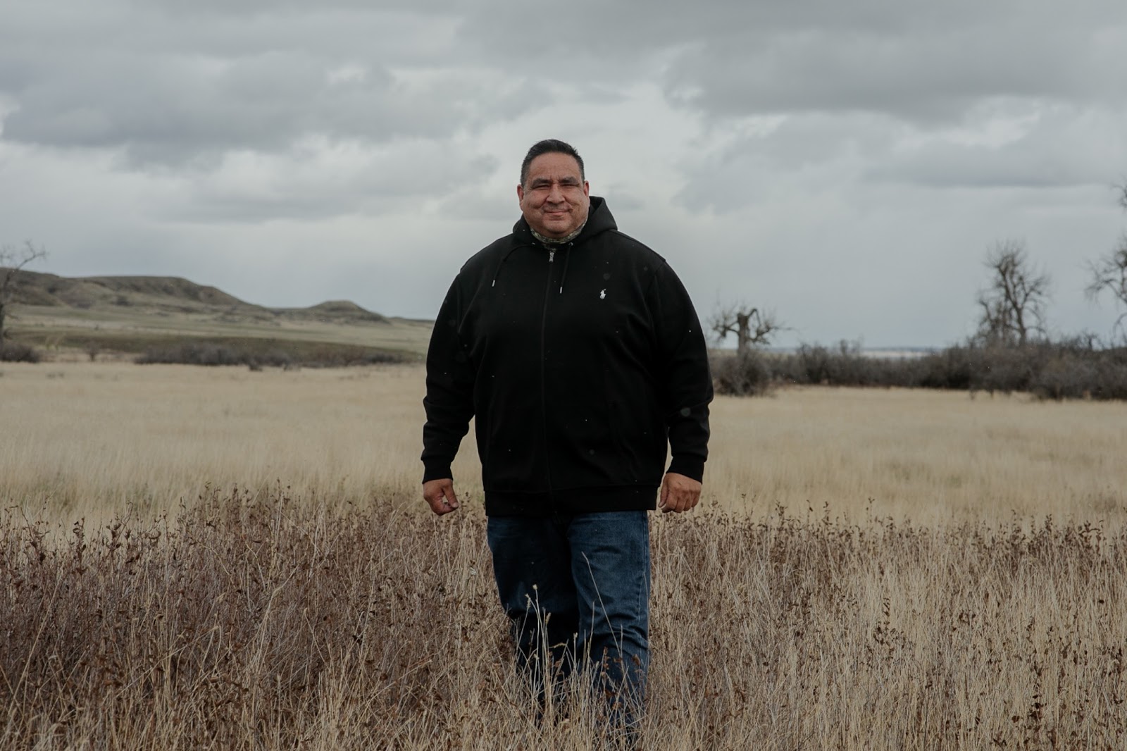 An Indigenous man in a black hoodie stands in a field against a cloudy sky