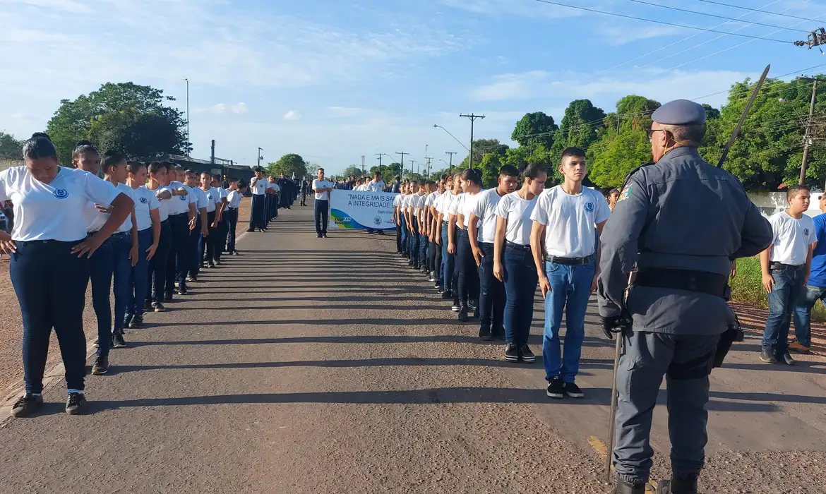 Três filas de jovens uniformizados parados com postura de atenção olhando para um oficial da policia. A cena acontece em ambiente externo