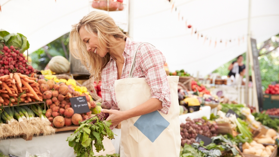 A smiling woman is shopping at a farmer's market, selecting fresh, local vegetables. Surrounded by colourful stalls filled with carrots, beets, zucchini, and other seasonal produce, she enjoys the warm and friendly atmosphere of the market.