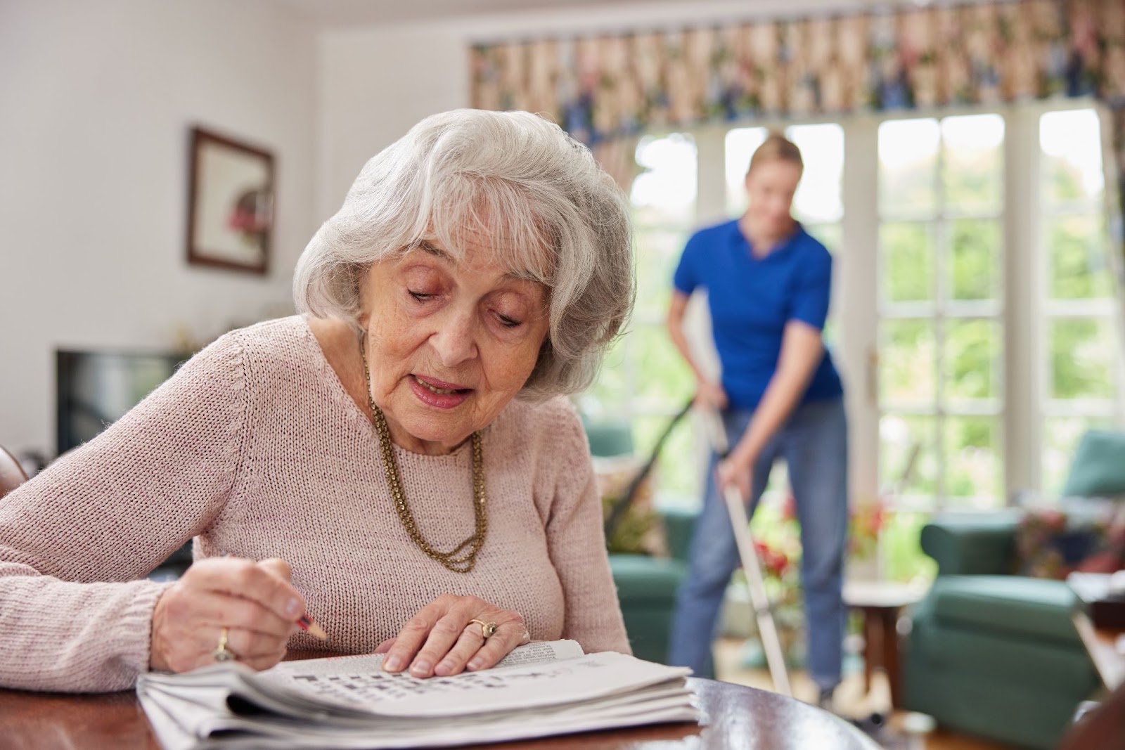 An elderly woman does the newspaper crossword as someone in the background helps clean her home in a Senior Living facility