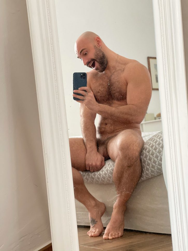 Mister Caccamo grinning with his mouth open naked sitting on the bed stroking his flaccid penis while taking an iphone mirror selfie for gay xxx porn photo content