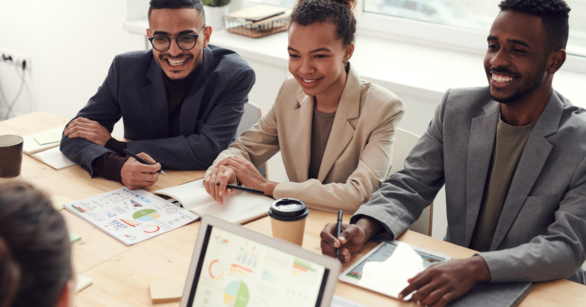 A diverse group of three professionals sitting together in a modern office, holding digital devices, and smiling confidently, showcasing the benefits of Scrum Master training.