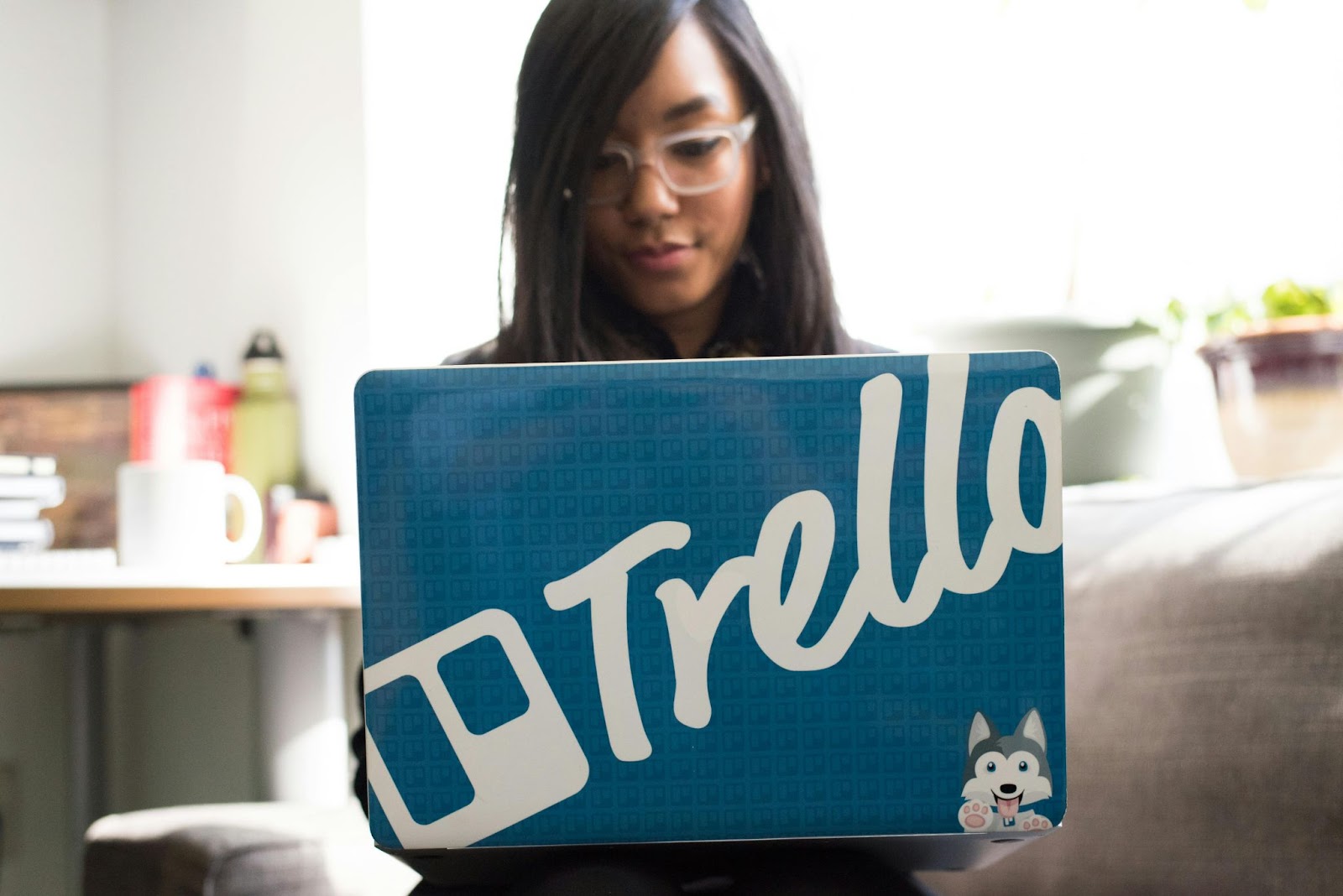 A woman using a laptop with a prominent Trello logo on the cover, working in a bright, casual workspace.