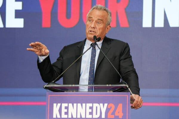 Robert F. Kennedy Jr. speaking at a lectern with a sign that says Kennedy 24. He is wearing a black sport coat and a skinny tie.