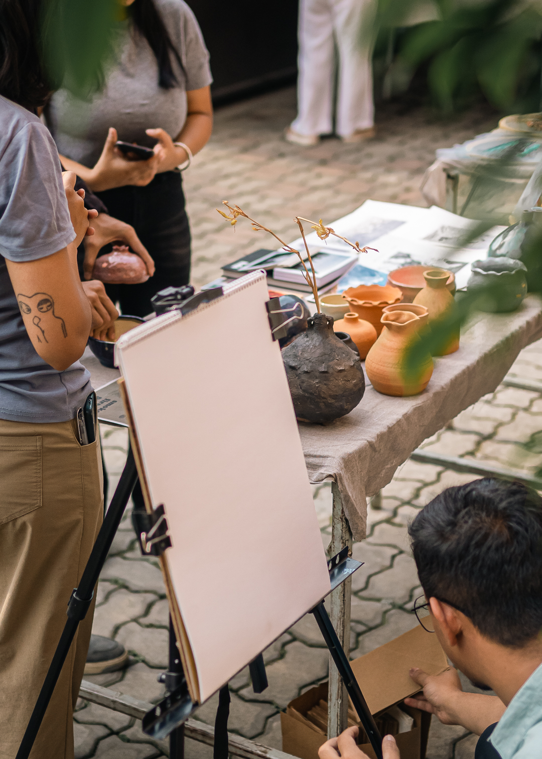 A table displays handmade ceramic pots and vases in various sizes and earth tones. An easel with a blank canvas stands in the foreground. Two people are partially visible: one with a tattoo on their arm and another using a smartphone. The scene is set on a stone-paved outdoor area, showcasing local artisans' sustainable crafts at the Nature's Market event. The composition emphasizes the blend of traditional craftsmanship with modern eco-conscious practices.