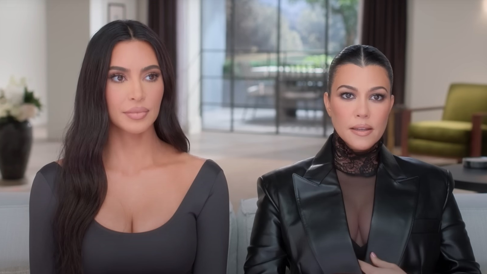 Kim Kardashian y Kourtney Kardashian sentadas juntas en un sofá, Kim lleva un top gris ajustado y Kourtney lleva un blazer de cuero negro con un top de encaje.