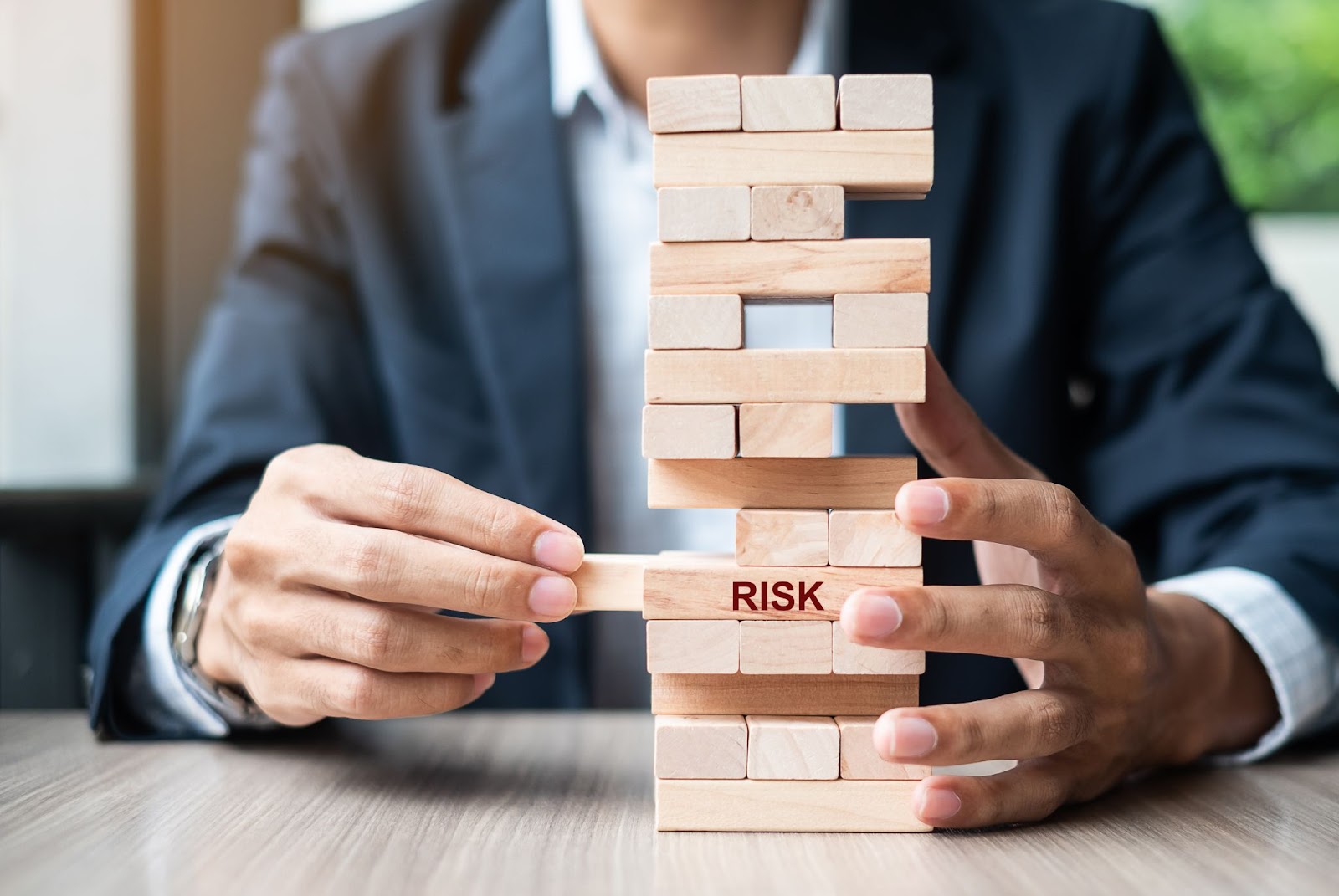 A businessman's hand in gray and white business attire is pulling a wooden block from a stack, symbolizing risk management’s impact. His face is not visible.