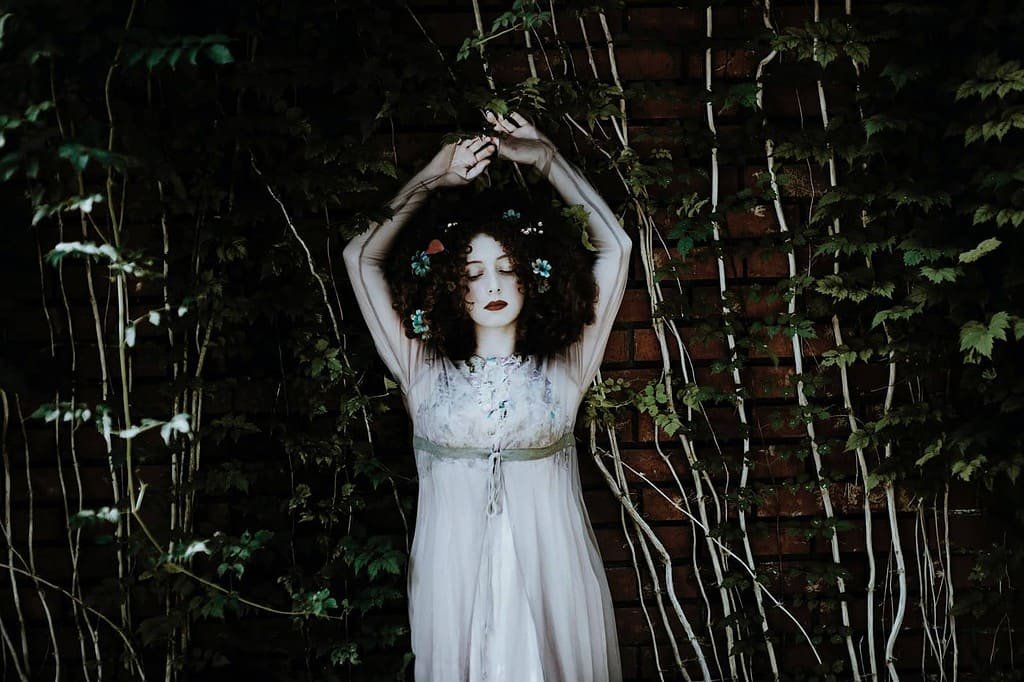 An individual stands against a dark background with dense foliage. They are wearing a white dress with floral patterns. The person’s arms are raised above their head, with hands touching, suggesting a pose or gesture. The image has an artistic composition with a contrast between the white dress and the dark background, adding a mysterious element.