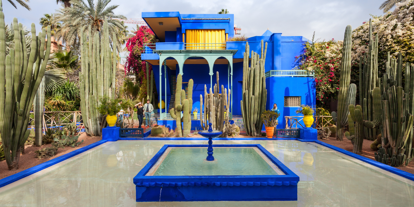 A vibrant scene of the Majorelle Garden in Marrakech, Morocco, featuring the iconic blue house surrounded by tall cacti and lush greenery. The garden's design includes a striking blue fountain in the foreground. Visitors are seen exploring the garden, highlighting its accessibility for travel enthusiasts. The combination of vivid colors and diverse plant life creates a captivating and serene atmosphere, perfect for accessible travel experiences.