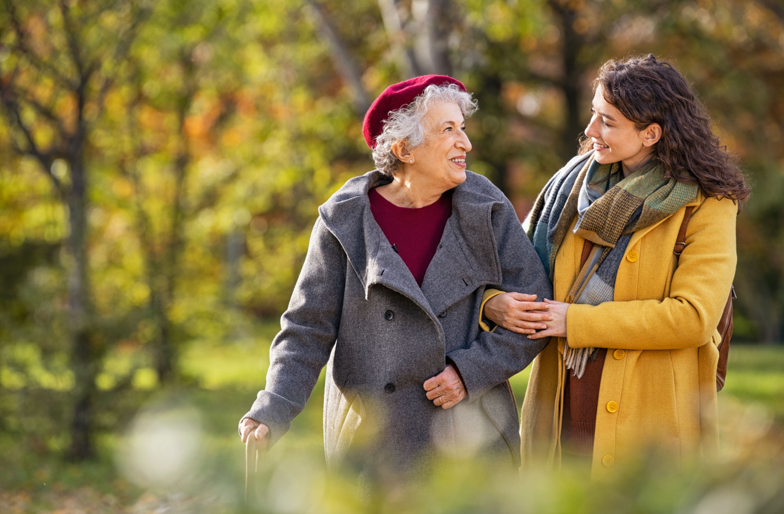 An adult child walks their senior parent through a bright, autumn-coloured park, smiling and holding each other.