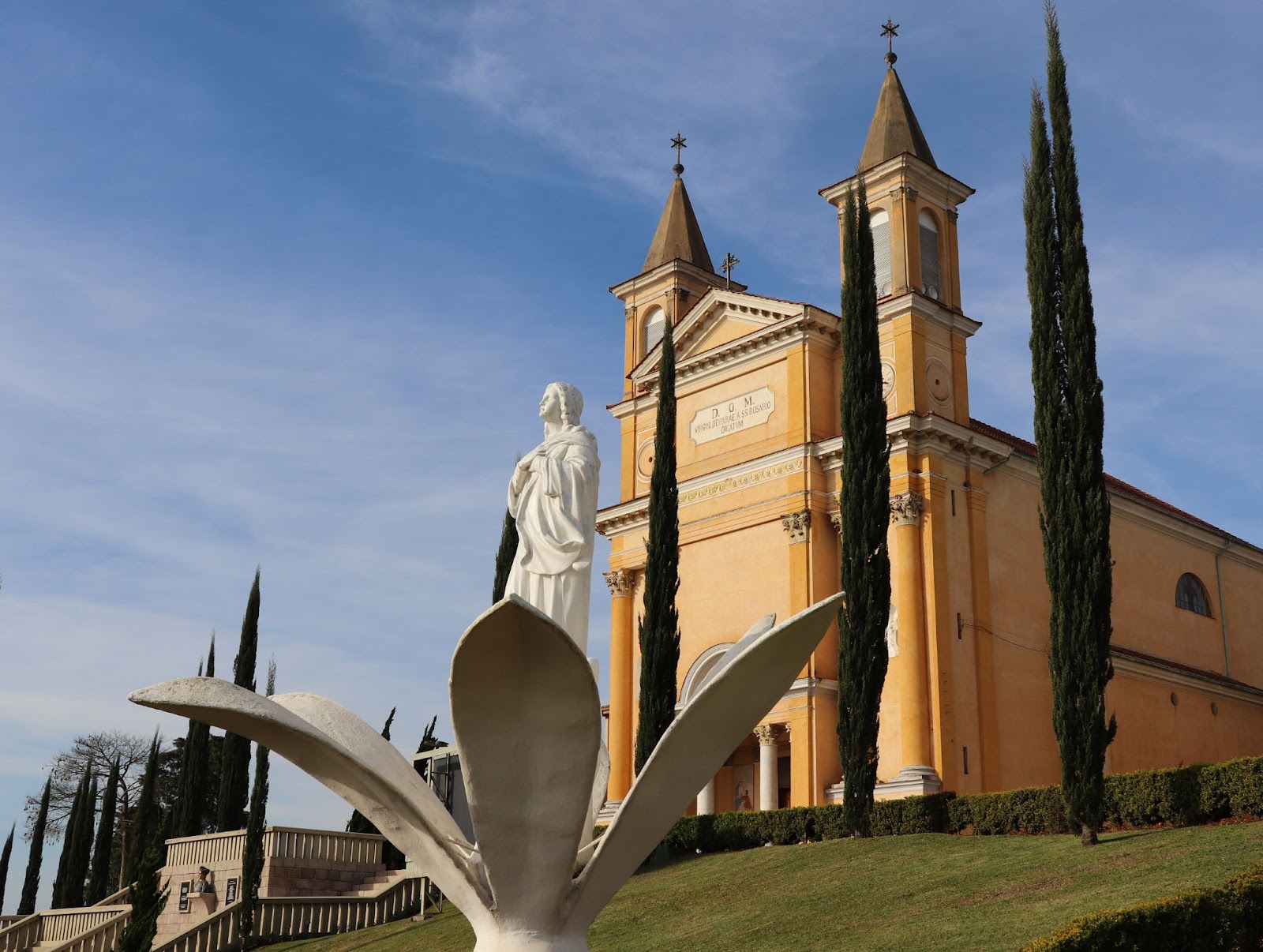 Igreja Matriz Nossa Senhora do Rosário ao lado direito da imagem, com estátua da santa à sua frente, e com foco na escultura de uma flor de lótus aberta.