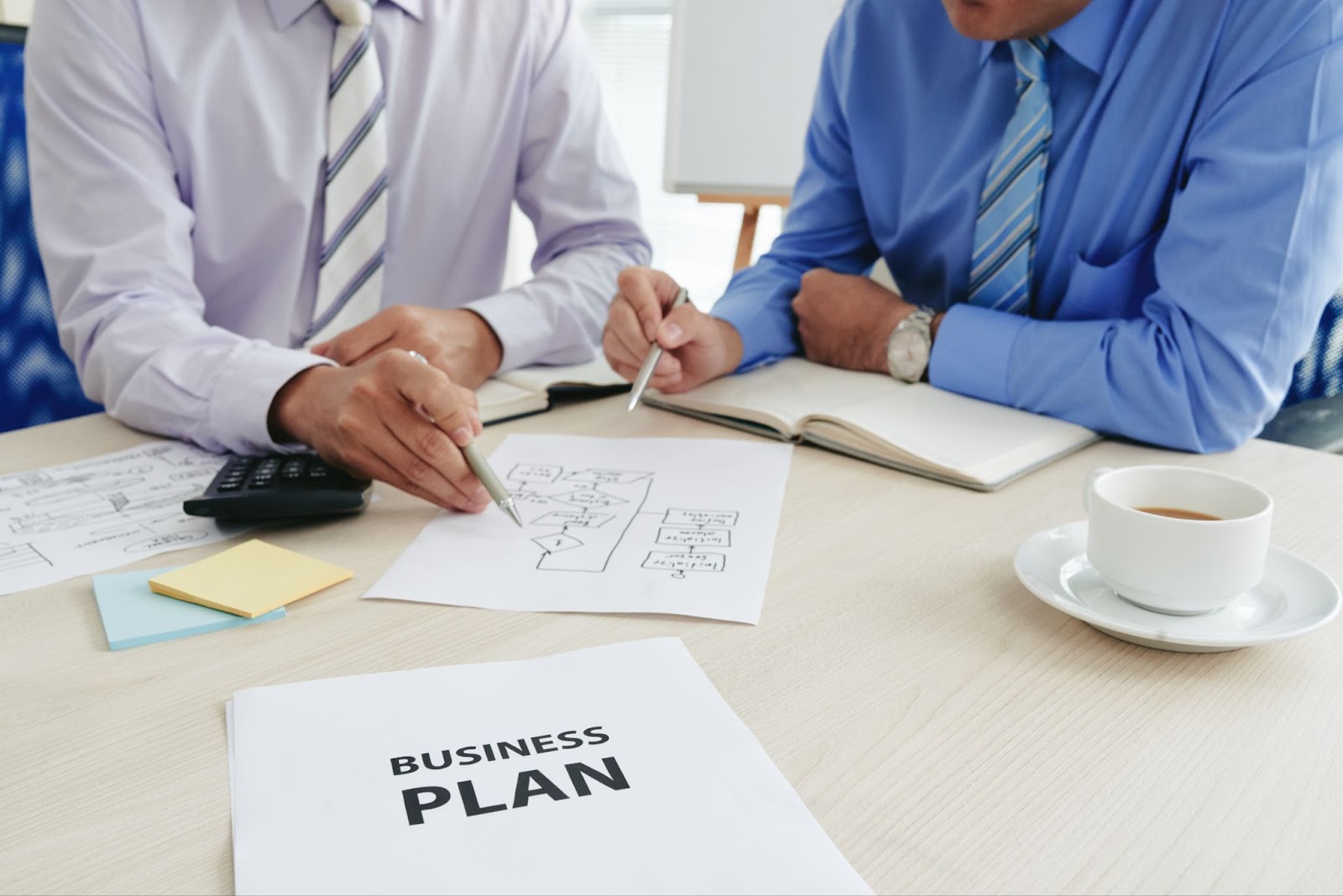  Two businessmen collaborating to create a business plan. They are seated at a table with a smartphone, a cup of coffee, and various documents spread out in front of them, deeply engaged in their discussion.