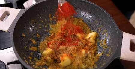 Various spices including turmeric, coriander, and Kashmiri chili powder being sprinkled over the sautéed potatoes and onion mixture.