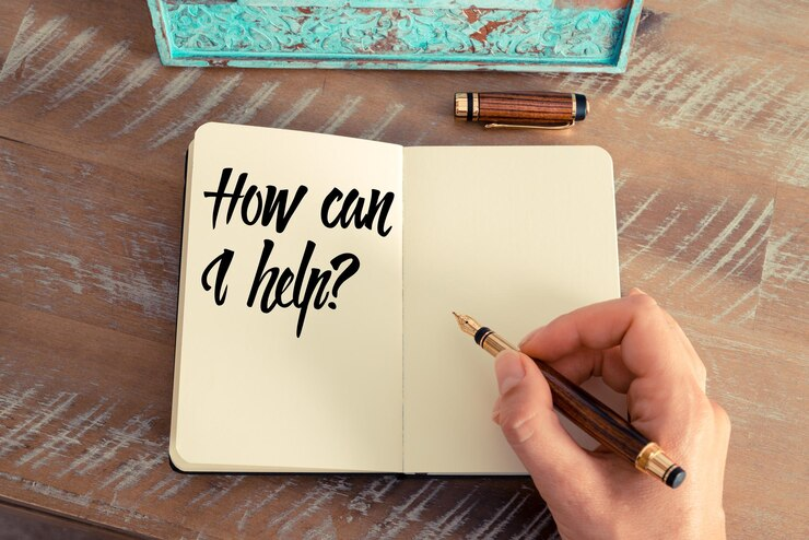 High angle view of hand holding a pen writing "How can I help?" on a notebook