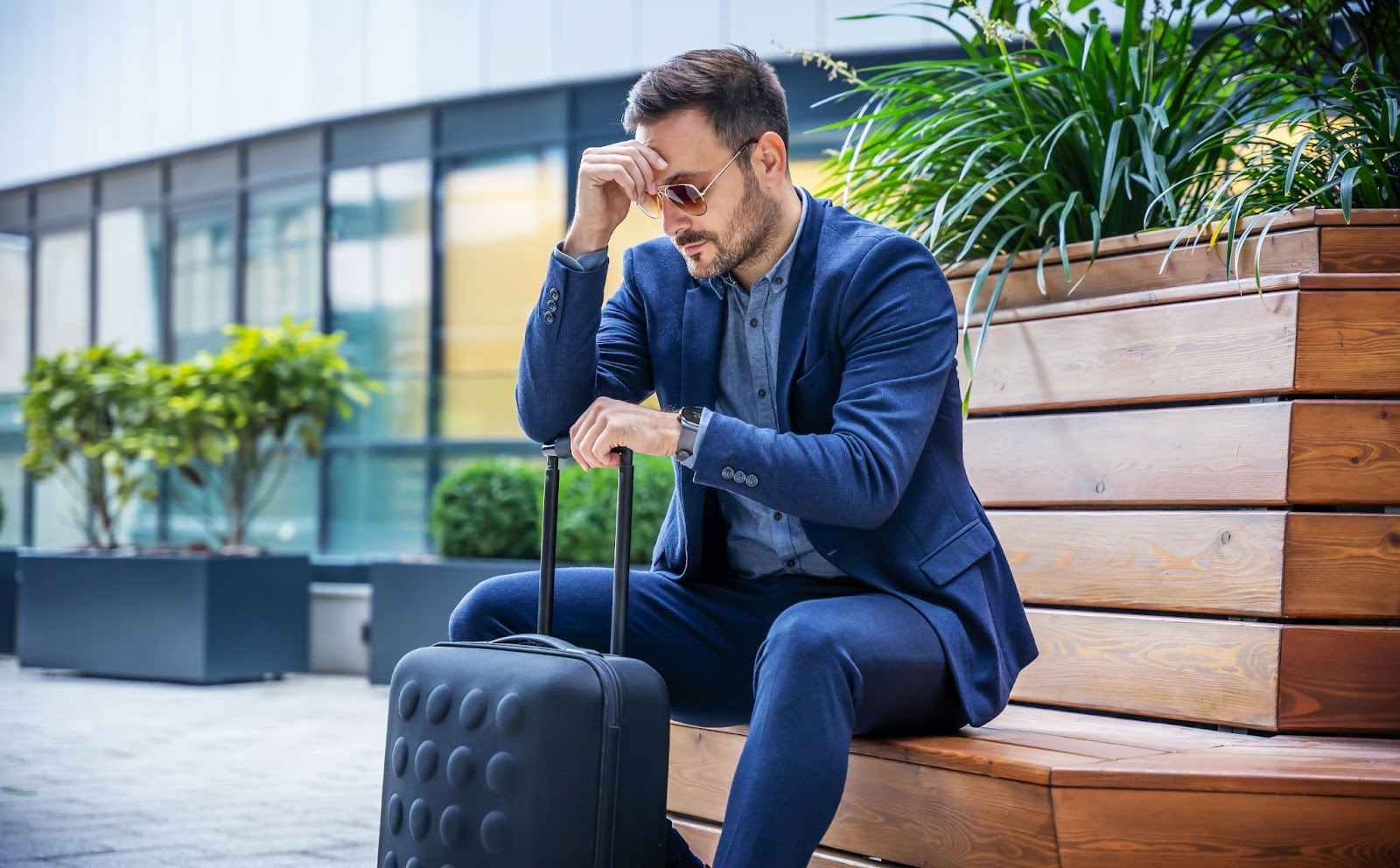 Homem, vestido socialmente, sentado em um banco de madeira, com uma mão no rosto, indicando exaustão, e outra mão na alça de sua mala de viagem.