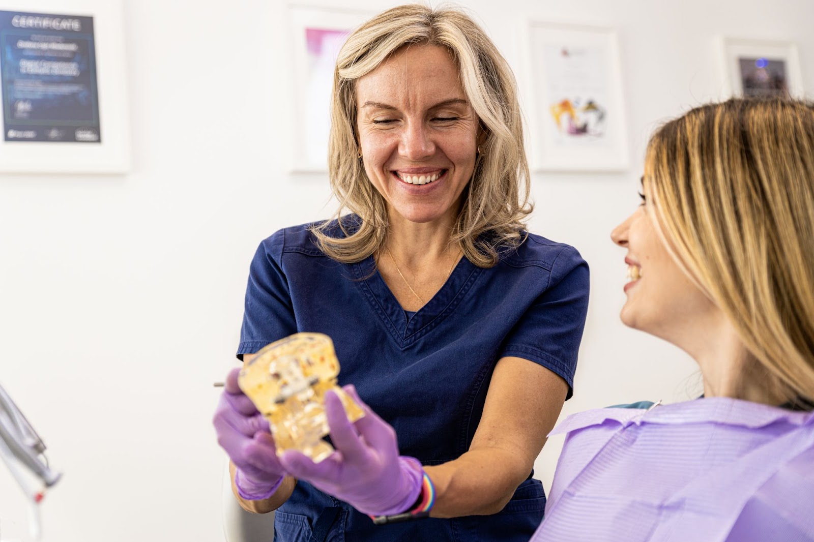 A female dentist wearing navy blue scrubs and purple latex gloves talks with a patient while showing an anatomical human jaw in a clinic; several picture frames are visible on the wall.