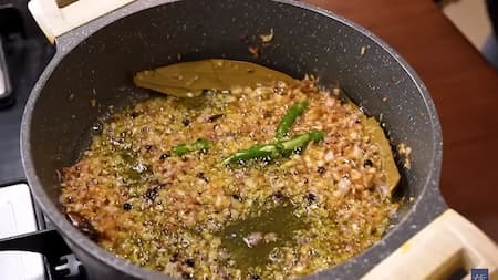 A pan with golden-brown onions, green chilies, and a mixture of whole spices such as bay leaves, black cardamom, and cloves, sautéing to create a rich masala base.