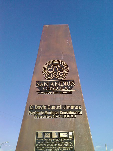 A tall, bronze-colored obelisk monument against a clear blue sky. The obelisk features engraved text in Spanish, commemorating the consolidation of a sports complex named Quetzalcoatl. 