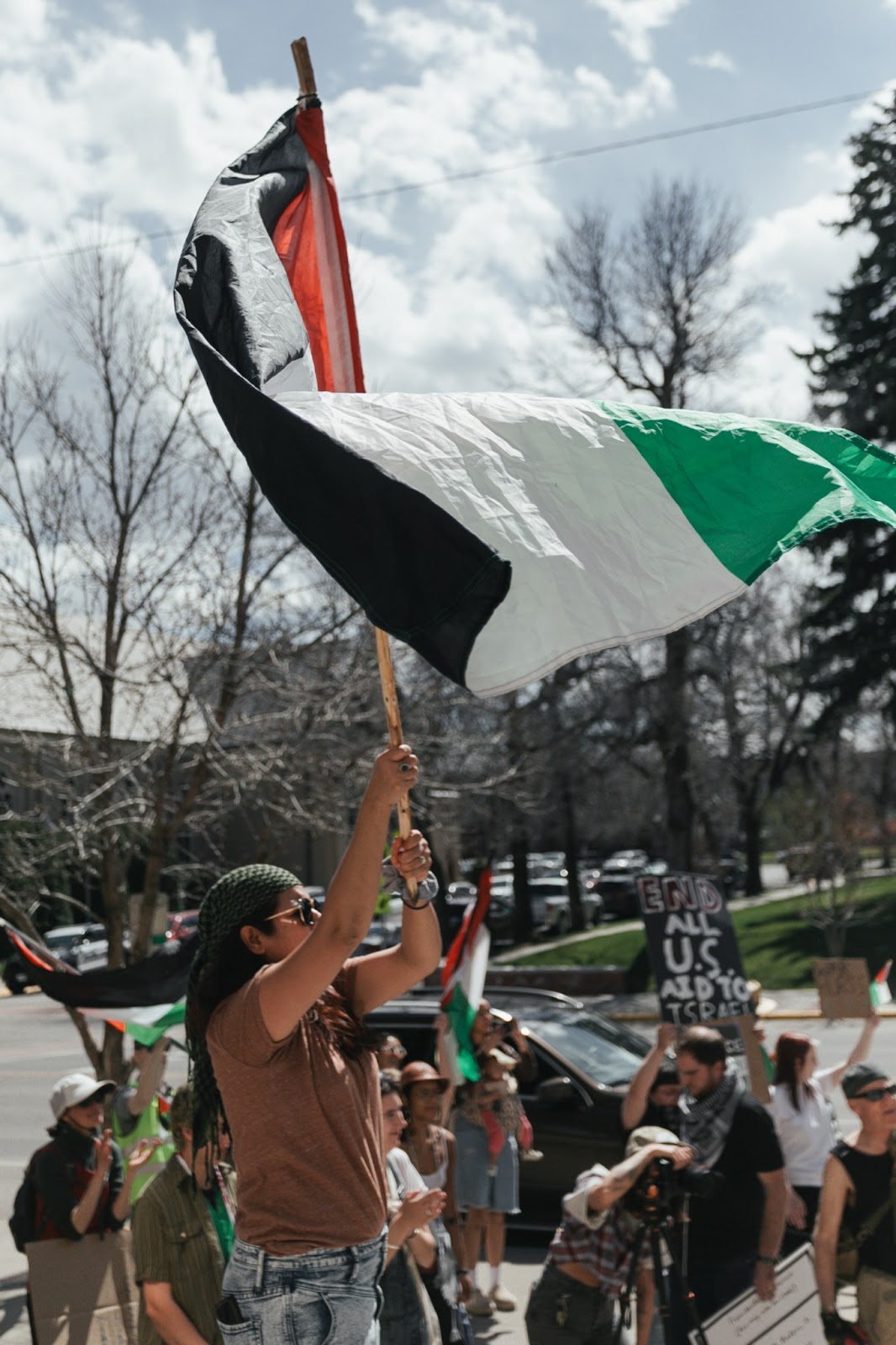 Color photograph of a person wearing a brown T-shirt and jeans waving a Palestinian flag in front of a small crowd. in the background, others also hold Palestinian flags