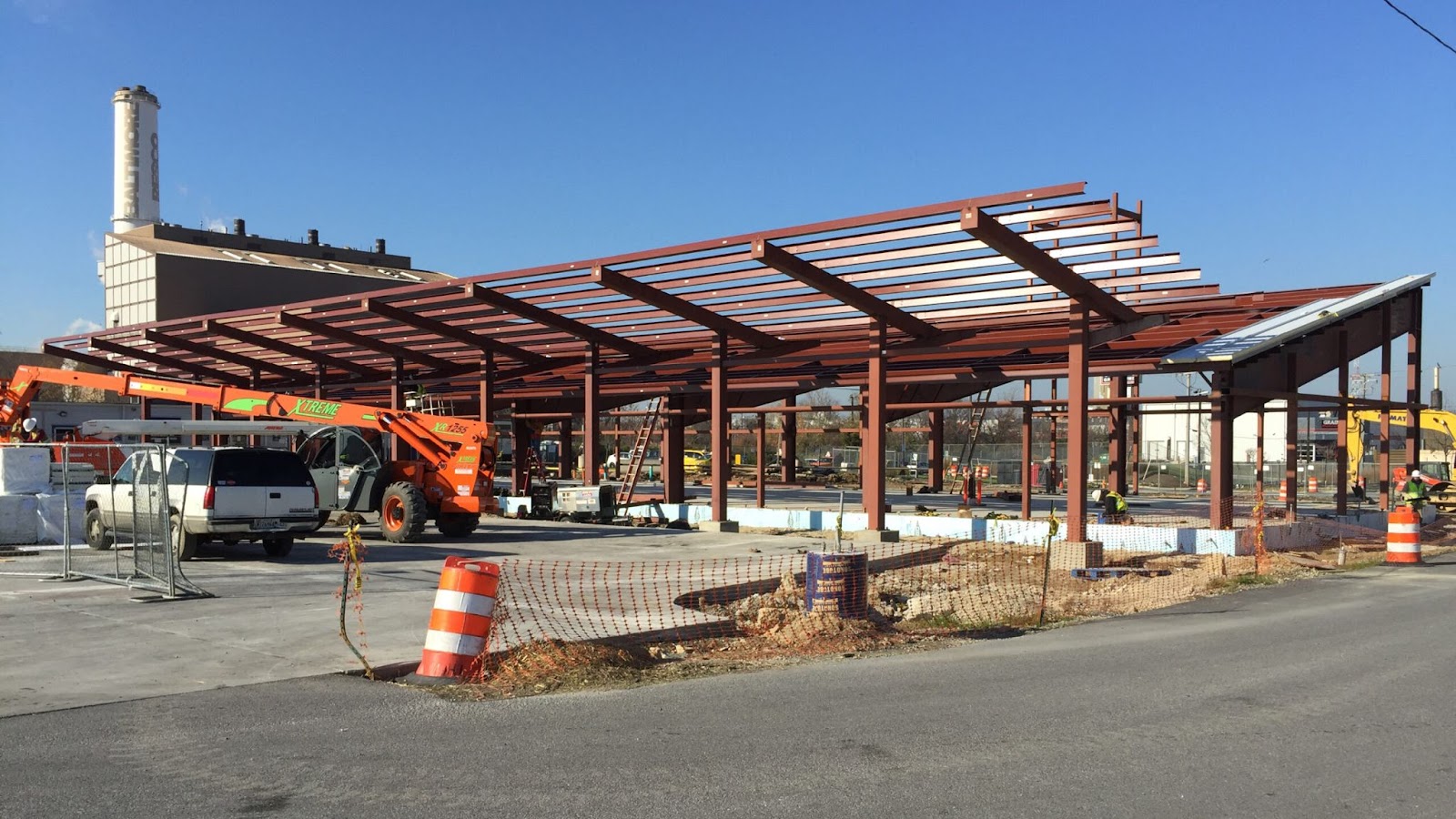 Construction site of a pre-engineered steel building with a visible steel framework. Construction vehicles and workers are present on the site, with an industrial facility in the background.