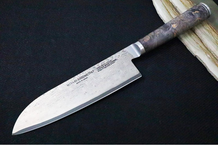 An image showing the Miyabi Black 7-inch Santoku knife on a solid black and wood background.