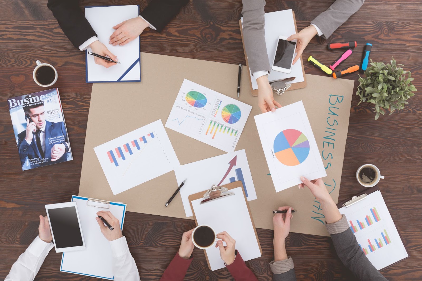 Top view of diverse business people collaborating around a table. They are working together to align business requirements with their strategic goals. There are various items on the table, such as a chart, a business magazine, a cup of coffee, a tablet, and colorful markers, all contributing to their productive meeting.