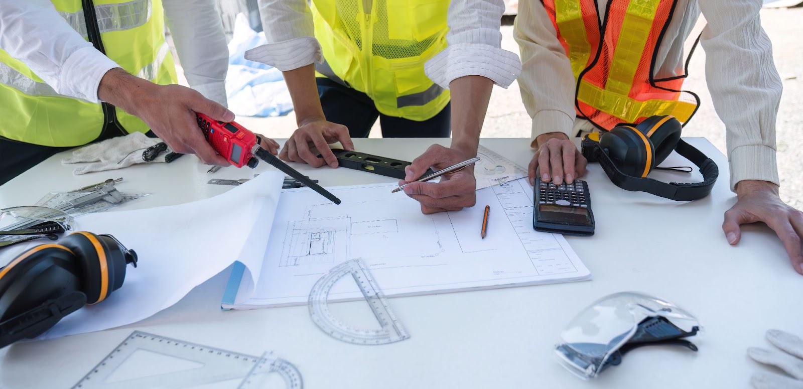 A cropped view of three construction workers’ hands using a calculator, a pen, and measuring tools to calculate costs.