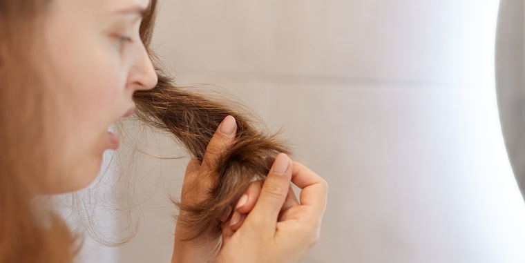 Imagem de uma mulher, vista de lado, enquanto segura com as mãos as pontas de seu cabelo e as observa descontente. Ela está localizada dentro de um banheiro em frente a um espelho.