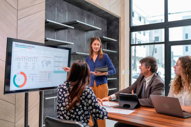 Businesswoman giving a data analysis presentation to colleagues in a modern office setting, using a digital screen to display charts and graphs.