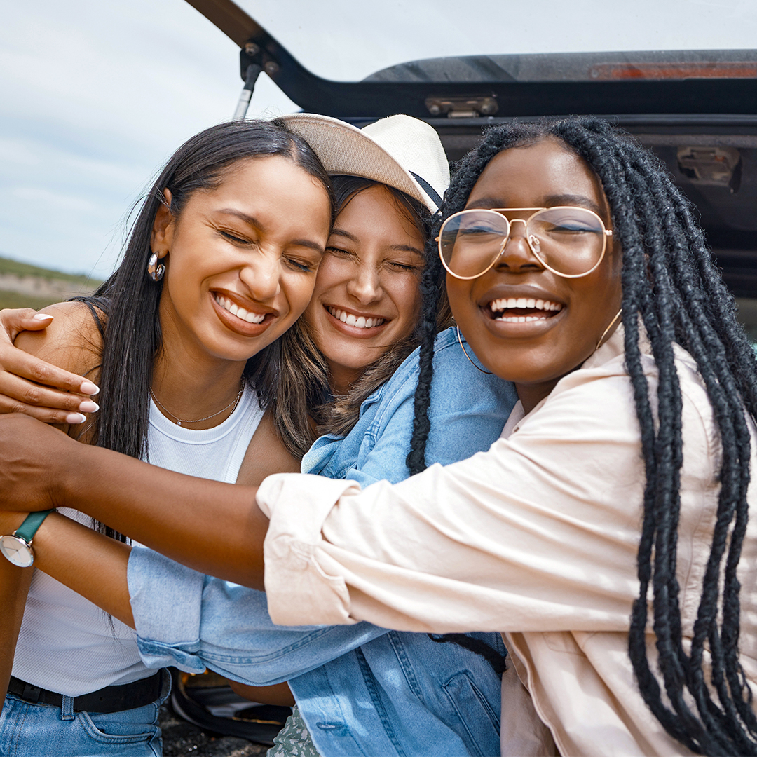 Three happy Gen-Z girl friends sit in the open trunk of their car on a sunny day, enjoying their road trip in New Mexico.