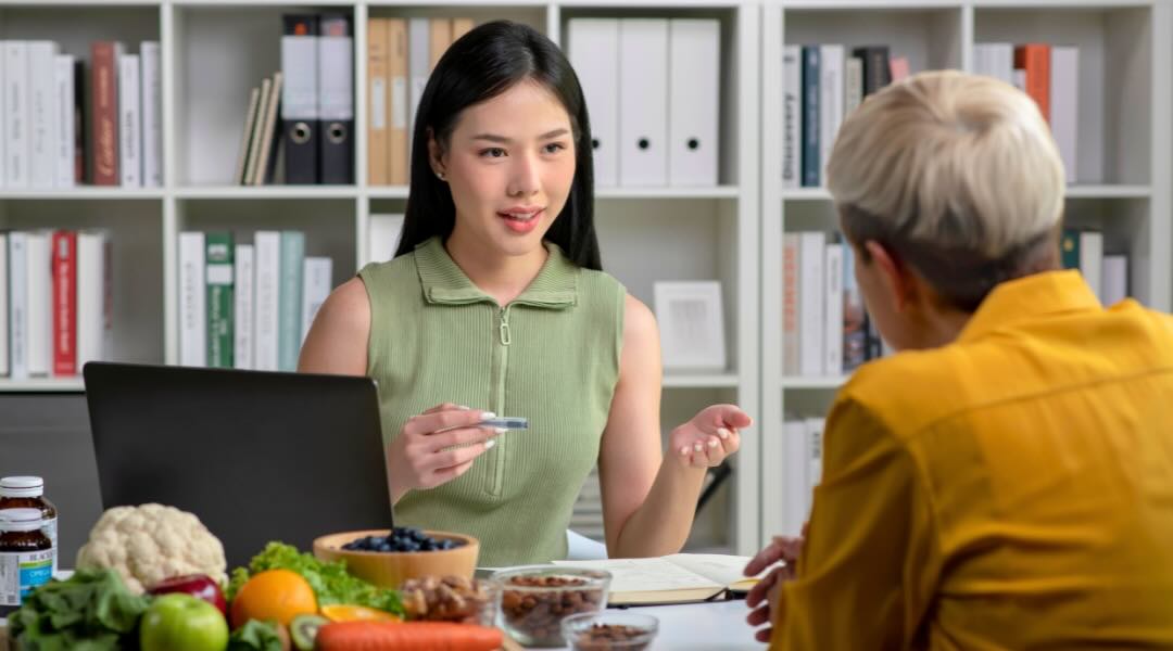 A female dietitian nutritionist sitting at a desk, engaging in a discussion with a client. The desk is adorned with various fruits and vegetables, and the background features shelves with books and binders.