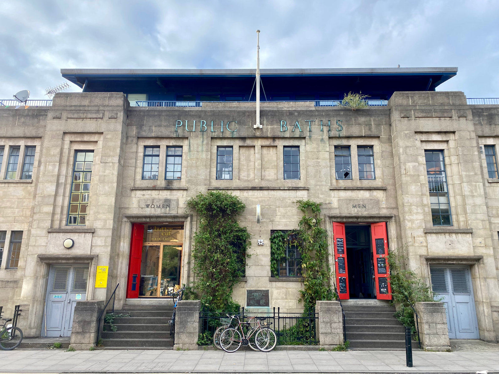 The old Public Baths building in Hackney Wick where the grief saunas are held. The building is large, square, and grey with red open doors.