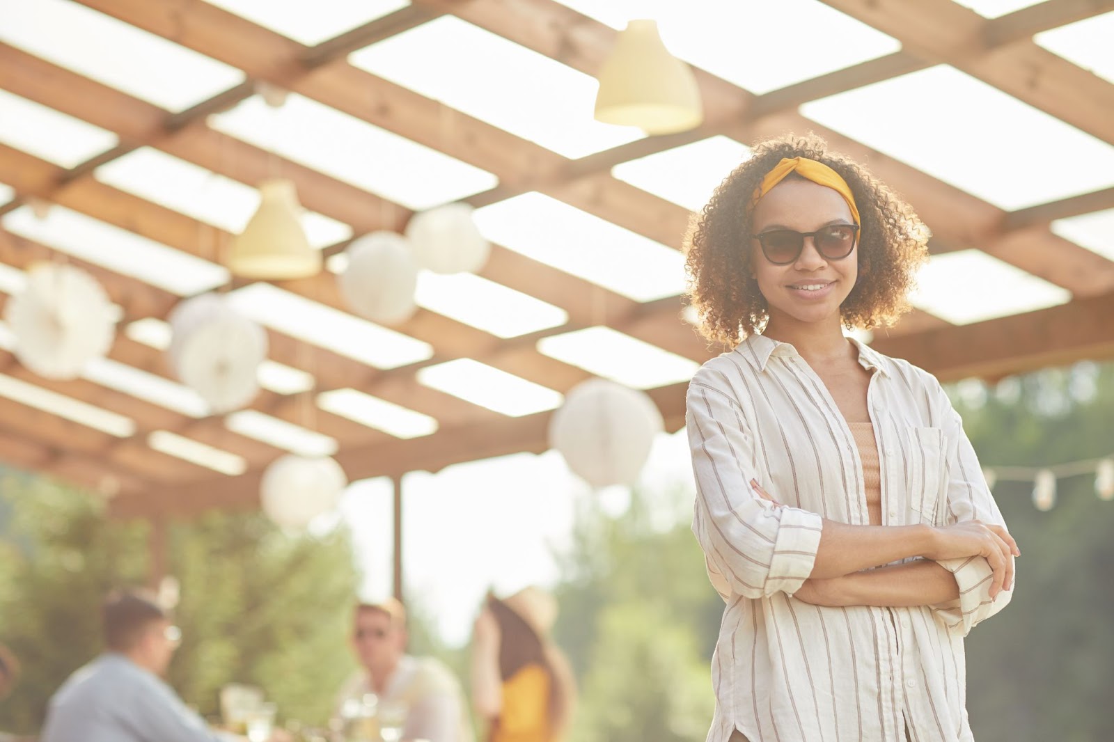 Woman wearing a yellow headband and sunglasses poses under a vinyl-covered patio with a blurred background of her friends enjoying drinks and conversation.