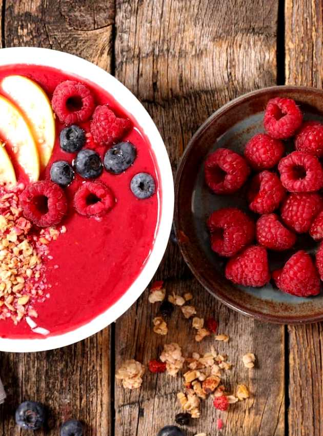 Red raspberry, banana, blueberry, and granola smoothie bowl on a rustic wooden tray.