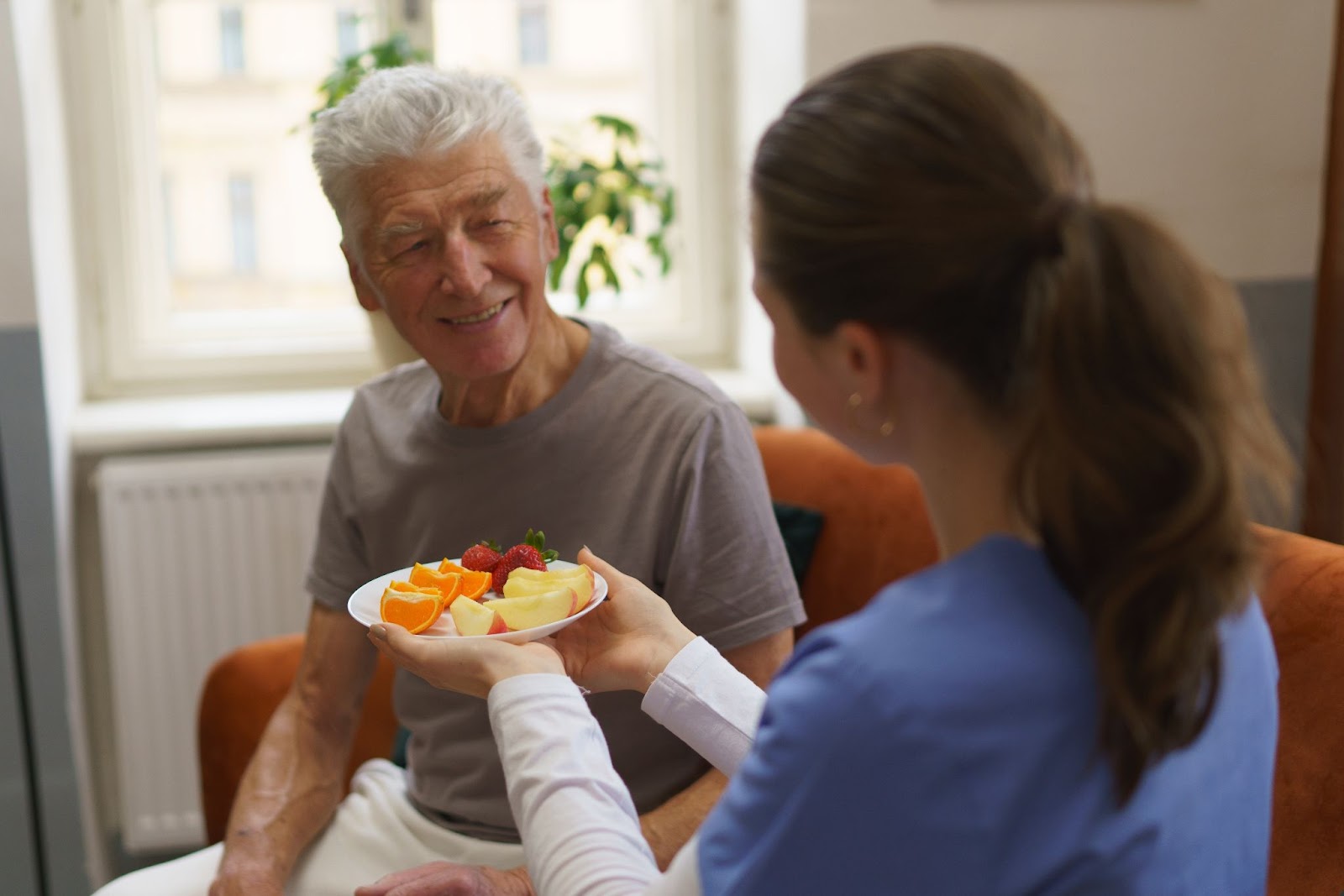 A young nurse handing a smiling senior man a plate full of sliced assorted fruits.