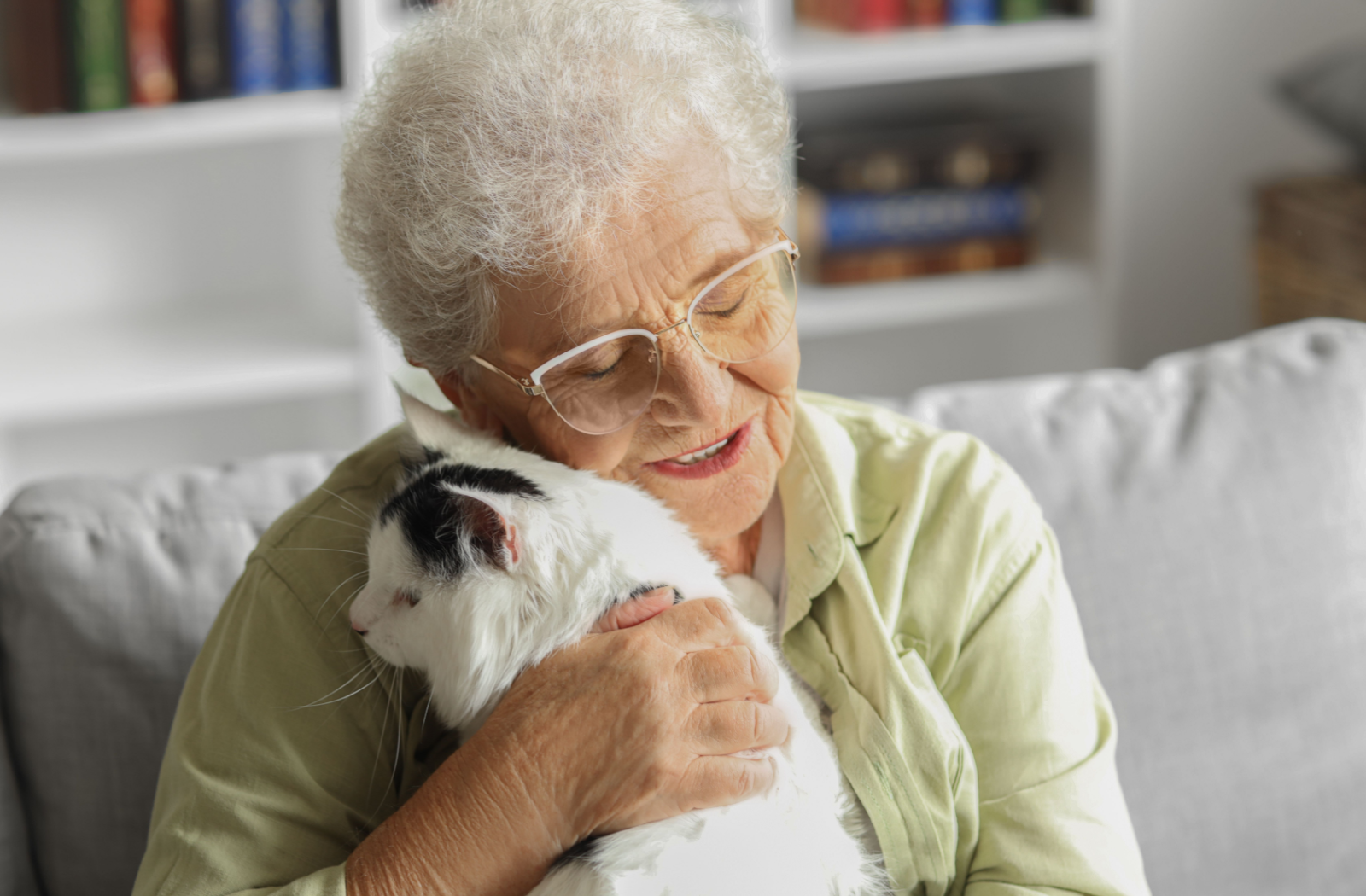 A senior sits on a plush, grey couch snuggling a black and white fluffy cat.