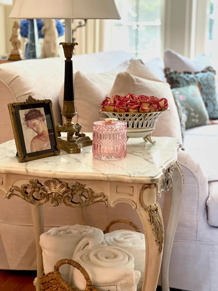 Ornate side table with marble top. Decor includes classic lamp, framed portrait, dried roses bowl, pink glass jar, and wicker basket with rolled towels.