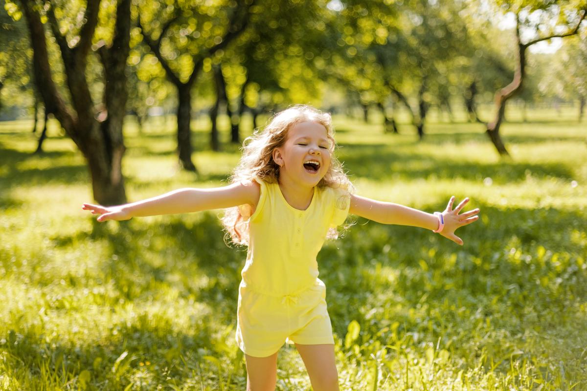 photo of a girl in a yellow dress with outstretched arms, in a field with trees on a summers day