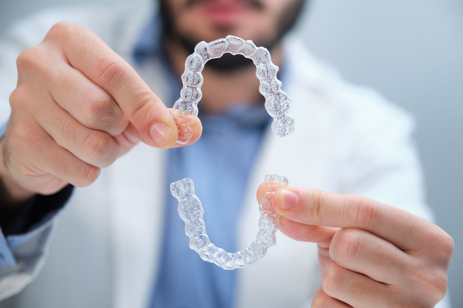 An unrecognizable bearded orthodontist holding clear trainer dental braces.