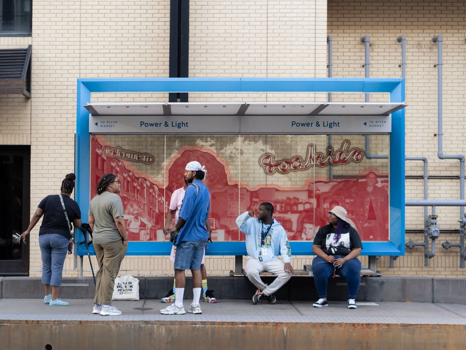 Image: A red-toned, collaged art installation depicting former Kansas City Black gay bar, Soakie’s, on the KC Streetcar Power & Light Northbound Stop––co-created with Zach Frazier. Montalvo, Taylor, Carr, and friends gather around the installation. Photo by Zach Frazier.