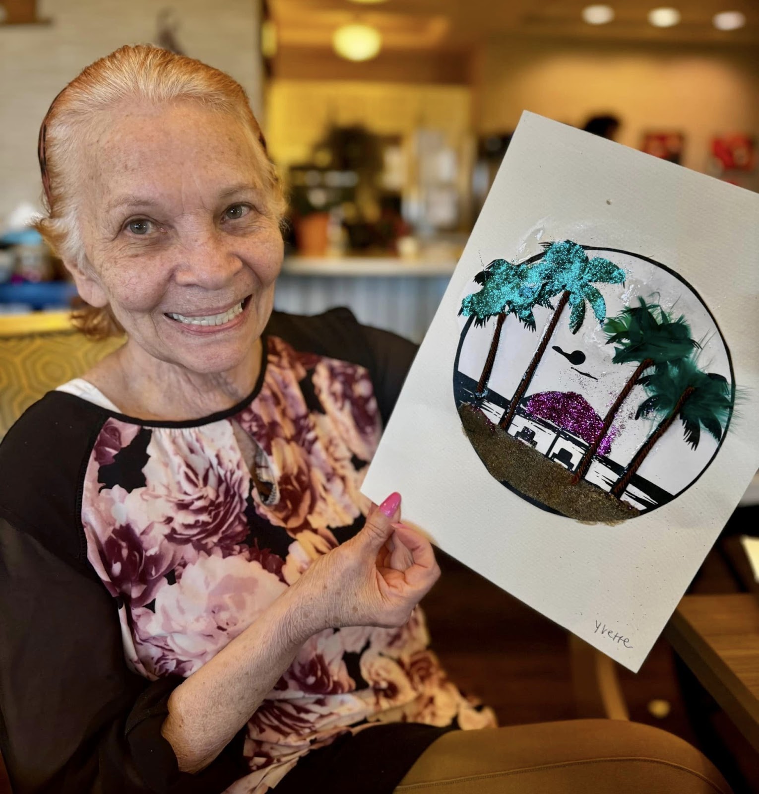 A woman smiling and showcasing a piece of art she created in an memory care/assisted living facility