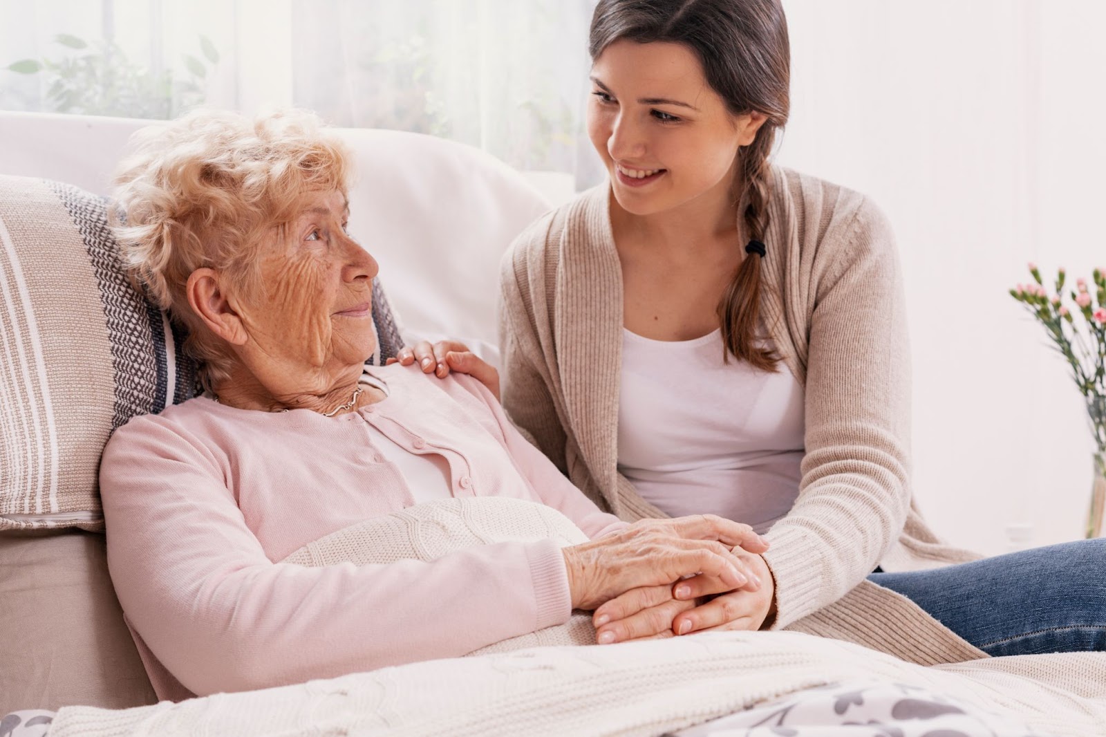 A caregiver gently supports a frail elderly woman who is lying in a hospital bed, offering comfort and assistance.