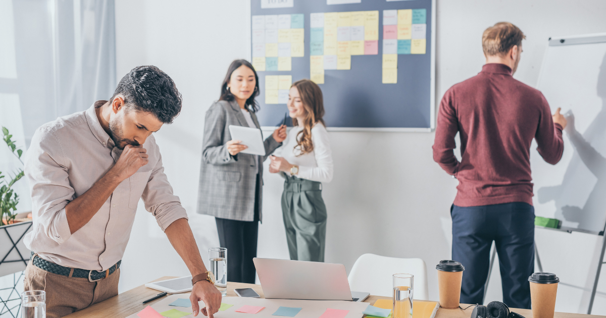 A diverse group of four professionals standing together in a modern office, holding digital devices, and writing on board, showcasing the benefits of Scrum Master training.