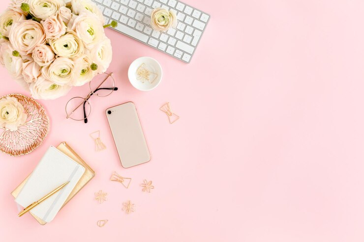 Stylized pink home office desk workspace with a keyboard, bouquet, phone, pair of glasses, and stationery on pink background