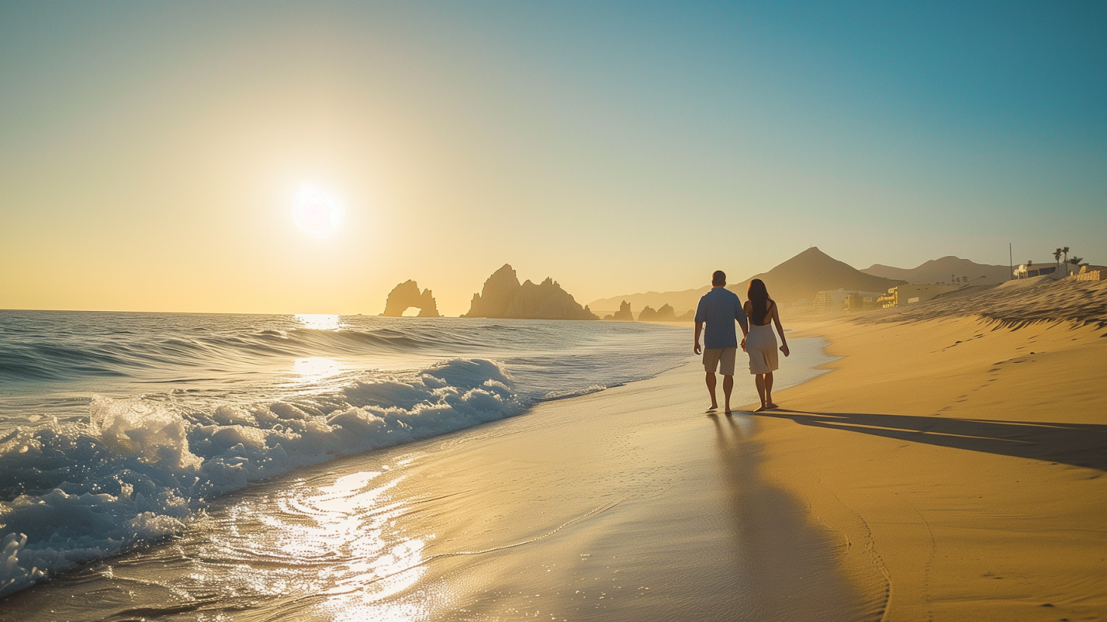 A couple walking hand in hand along Medano Beach with the Arch of Cabo San Lucas in the distance.