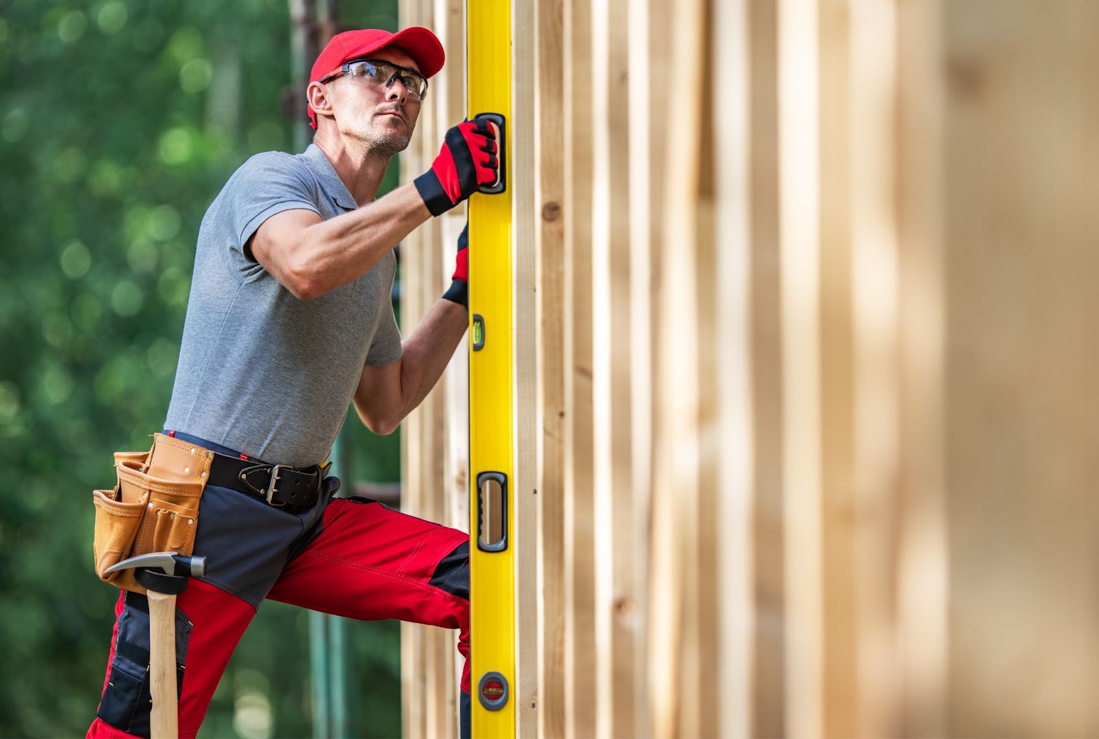 A Caucasian fence contractor in red and black safety gear and a gray shirt uses a bubble spirit level to measure a wooden fence for accuracy. A blurry green bush is seen in the background.