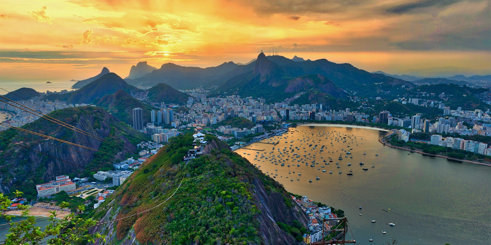 A breathtaking aerial view of Rio de Janeiro, Brazil, at sunset, with the city's iconic mountains and coastline. The sun casts a golden glow over the bay, dotted with boats, and the lush green hills contrast beautifully with the urban landscape. The famous Christ the Redeemer statue can be seen in the distance atop Corcovado Mountain, adding to the dramatic and picturesque scenery. The vibrant colors of the sky and the tranquil water create a serene and captivating atmosphere.